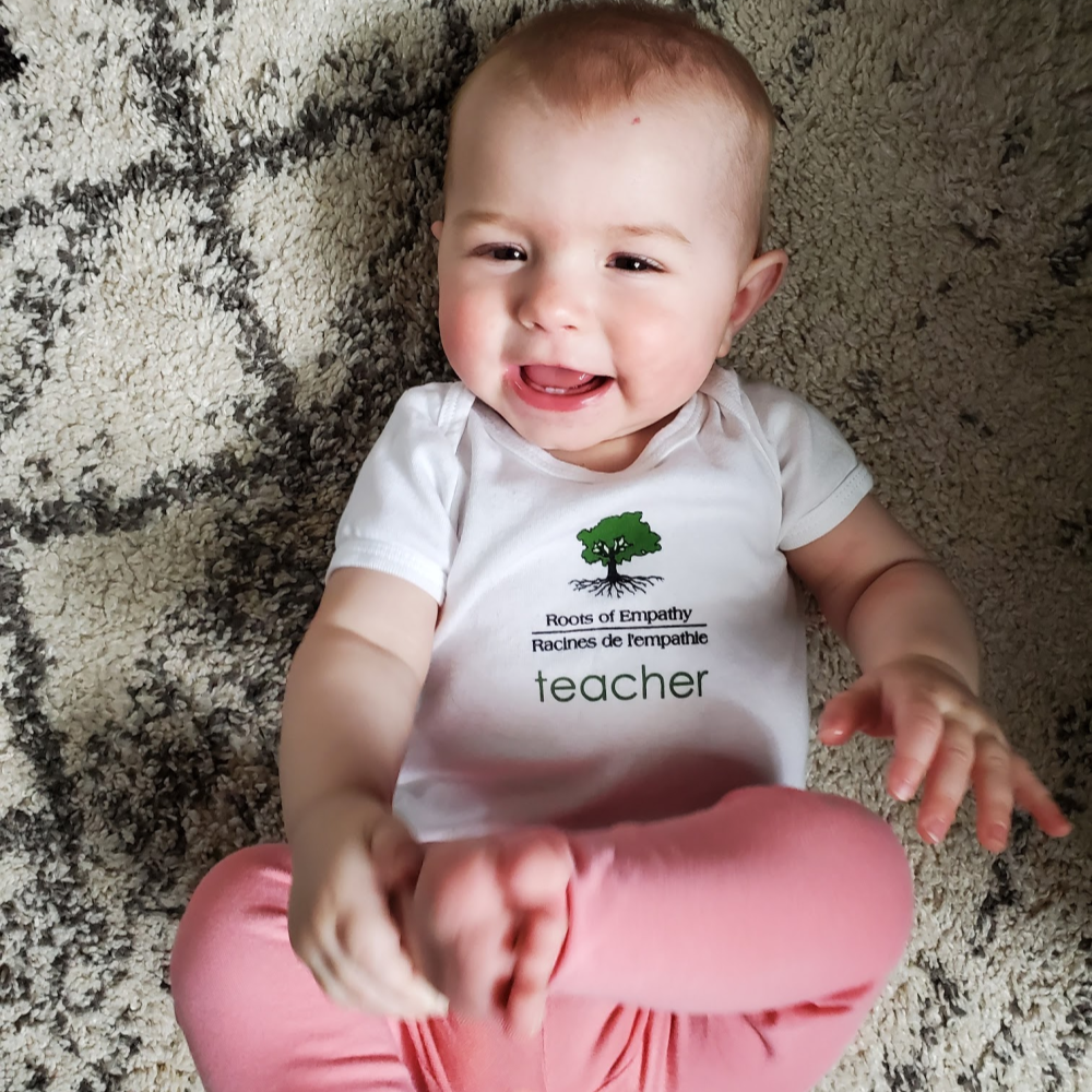 Baby Arden lays on her back on the rug, smiling at camera, wearing her teacher tshirt and pink leggings. She's playing with her foot.