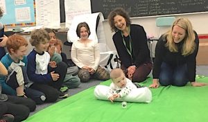 Baby on green blanket in a classroom. People sit around him watching him.